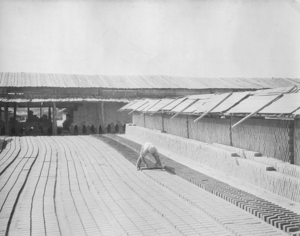Black and white photo of a person bent over many rows of bricks outside. Stacked bricks are seen on the right.