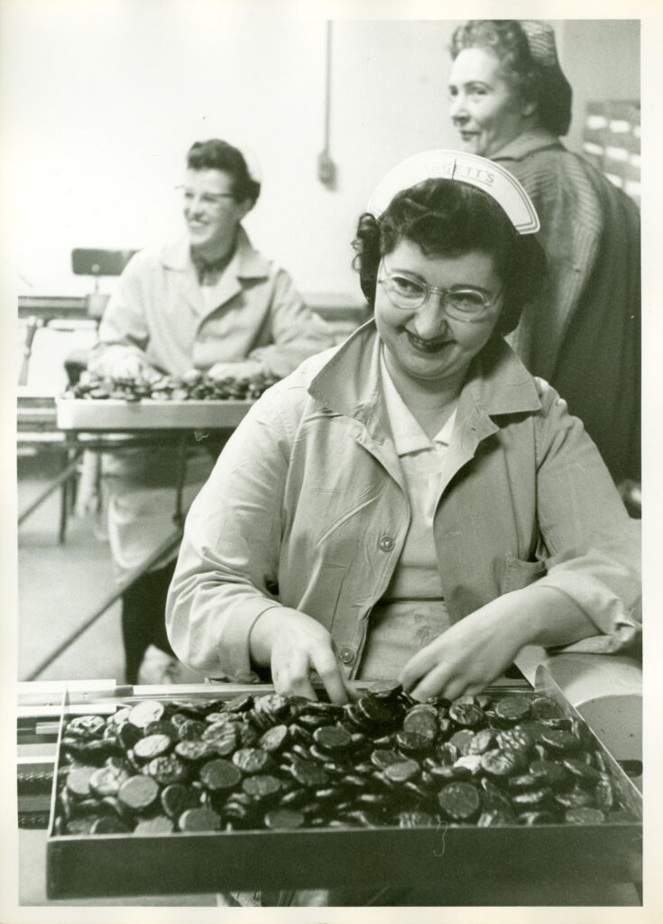 Black and white photo of a seated person with candy on a table in front of them. Two other workers can be seen in the background.