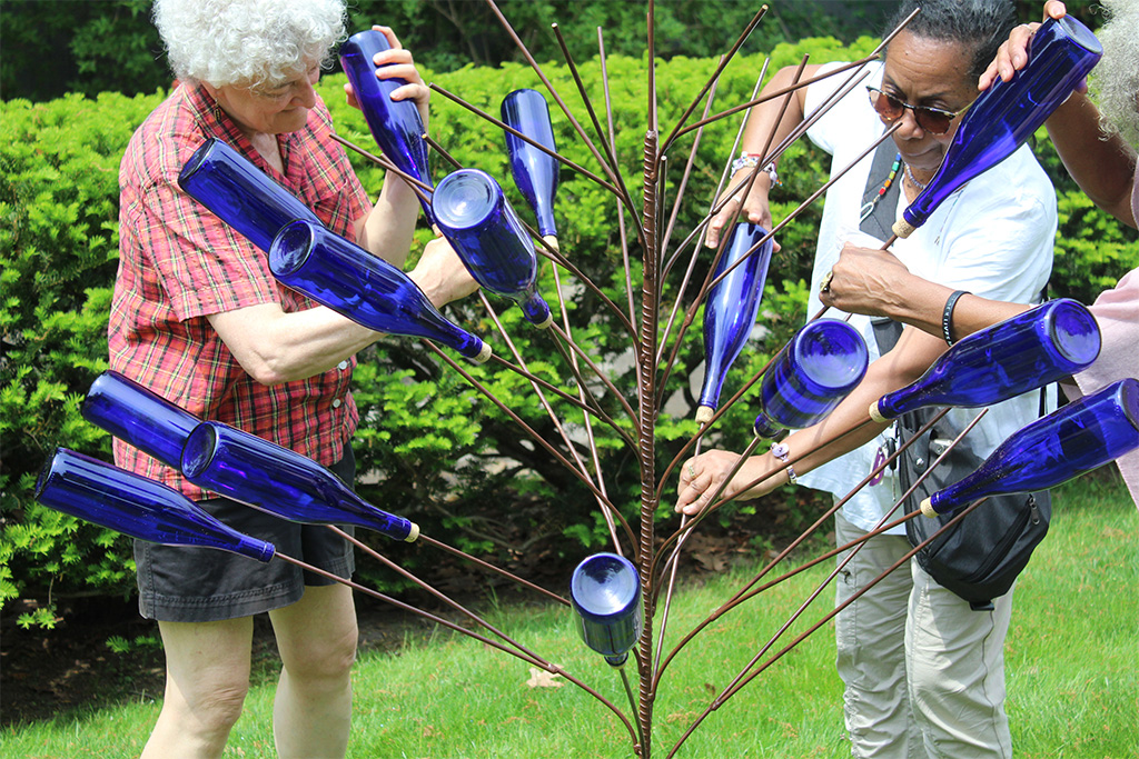 Two people screwing blue bottles onto a bottle tree in front of a hedge. Additional pair of hands on right.