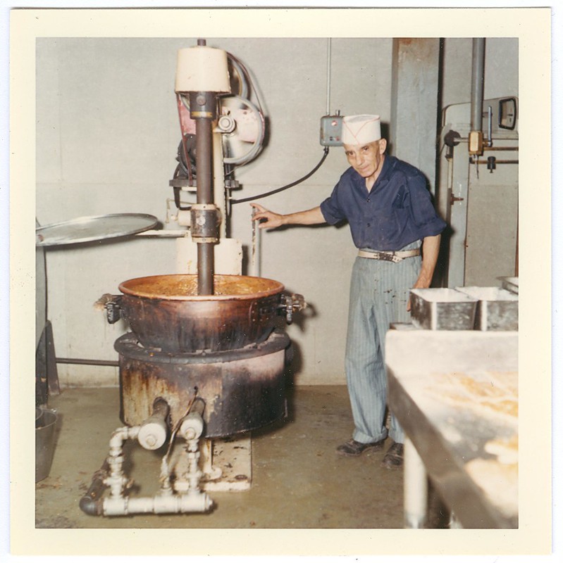 Colored photo of a man in a blue shirt and white hat touching a machine in a factory