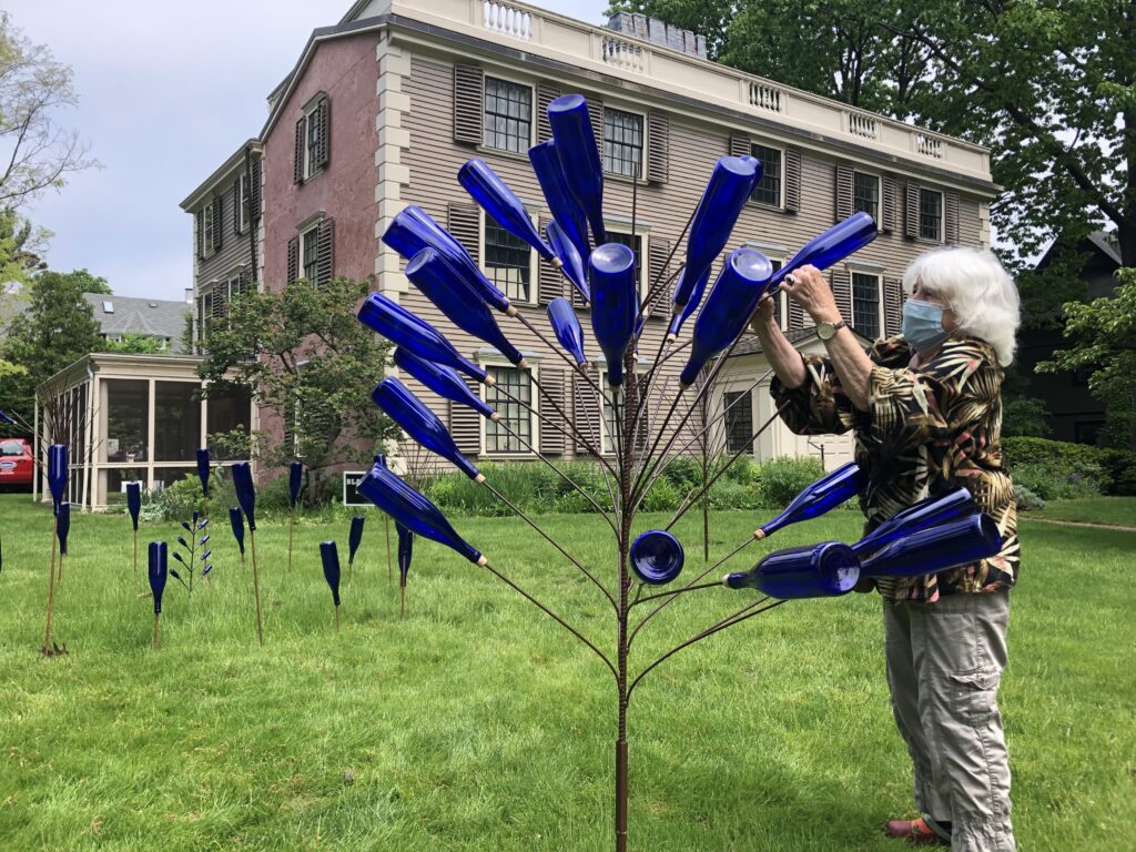 Person screwing a blue bottle on a bottle tree in front of a three story building with a green lawn.