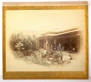 Black-and-white photograph with colored-in blue, red, yellow and green details representing four standing and sitting Japanese women and two seated white men at the rear of a Japanese house with a garden on the left.