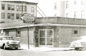 Street level view of one story brick building with a double door entrance. sign reads "Simeone's/ Italian American/ Restaurant/Pizza". Two cars parked curbside.