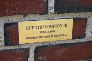 Close up of a shiny brass plaque on a red brick wall that reads: "Quentin Compson III

June 2, 1910

Drowned in the Fading of Honeysuckle"