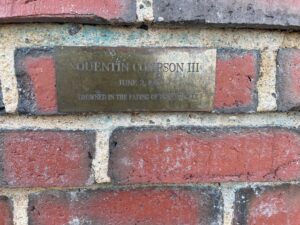 Section of a red brick wall. A rectangular brass plaque reads, "Quentin Compson III June 2, 1910 Drowned in the Fading of Honeysuckle"