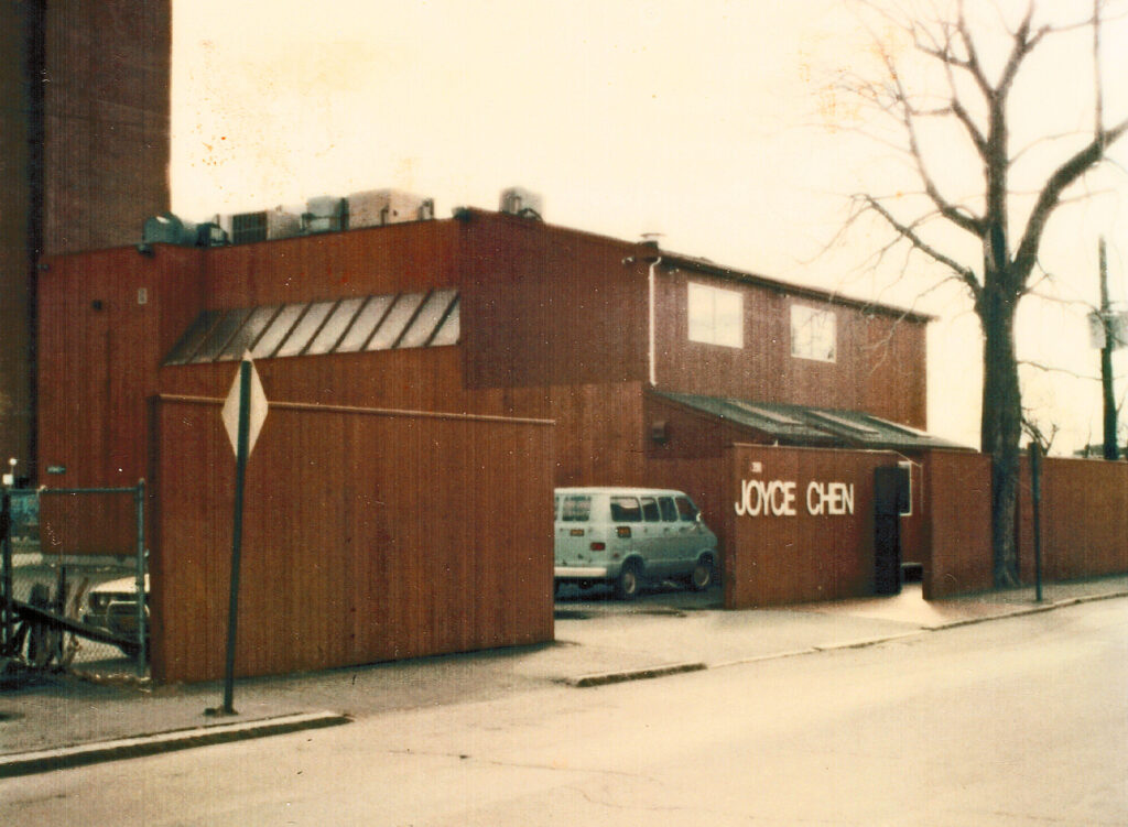 two-story brown building with "Joyce Chen on the side." A blue car is parked in back.