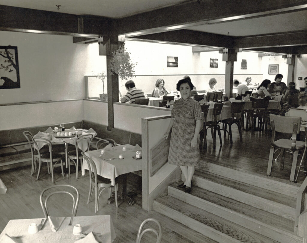 Woman standing on a short set of stairs inside a partially-full restaurant.