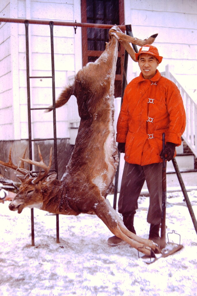Man standing next to a deer hung by its hind legs from a children's swingset in the snow.