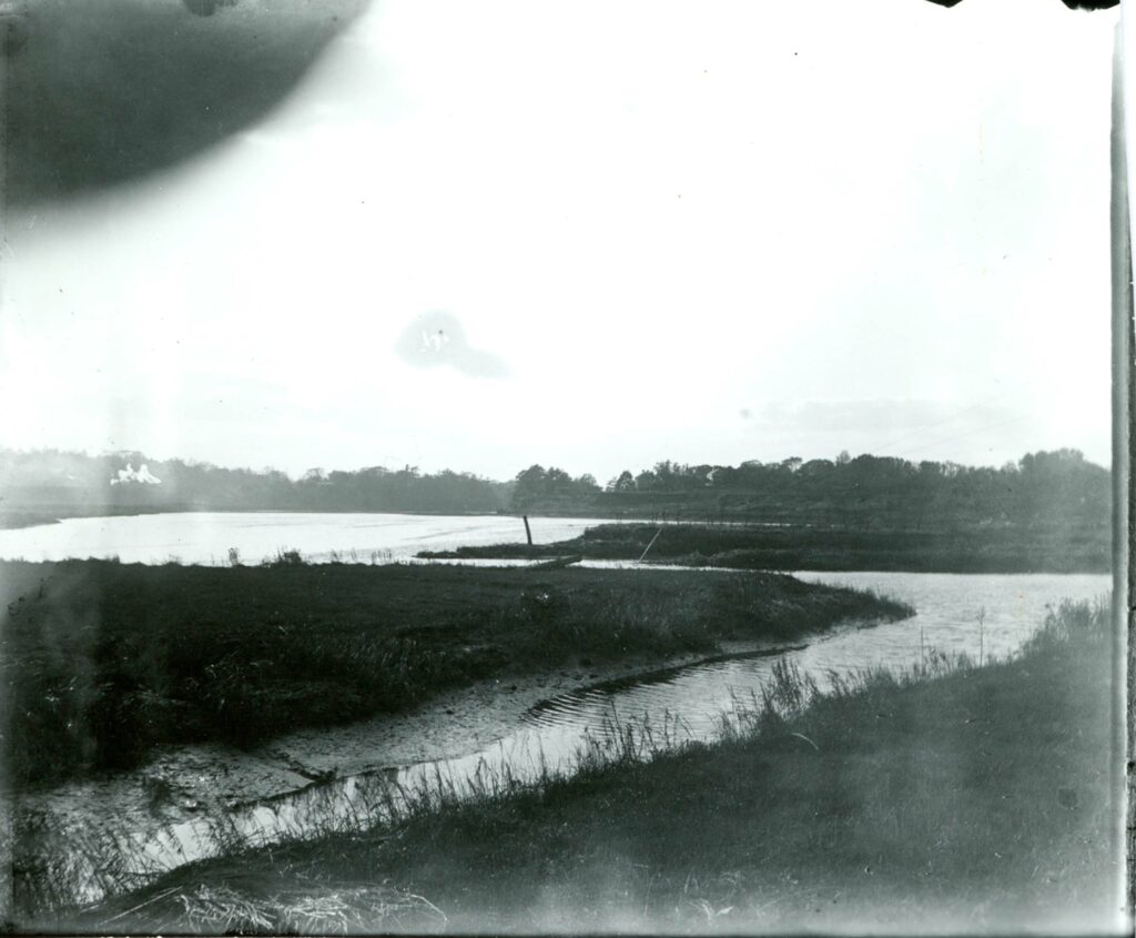 Black and white photo of a winding stream. Trees on the bank in the distance.