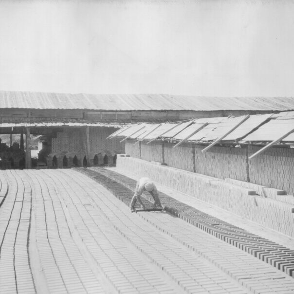 Black and white photo of a person bent over many rows of bricks outside. Stacked bricks are seen on the right.